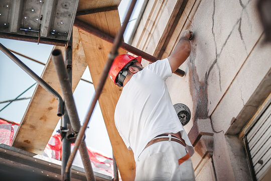 Mann mit rotem Helm steht auf einem Außengerüst und bearbeitet die Außenwand eines Gebäudes.