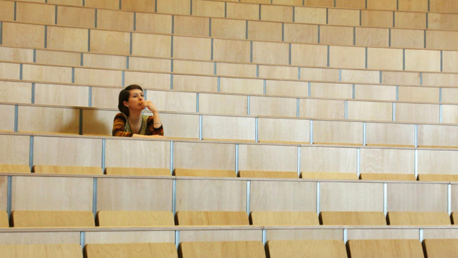 Eine junge Person sitzt alleine in einem Hörsaal und blickt nachdenklich nach vorne.