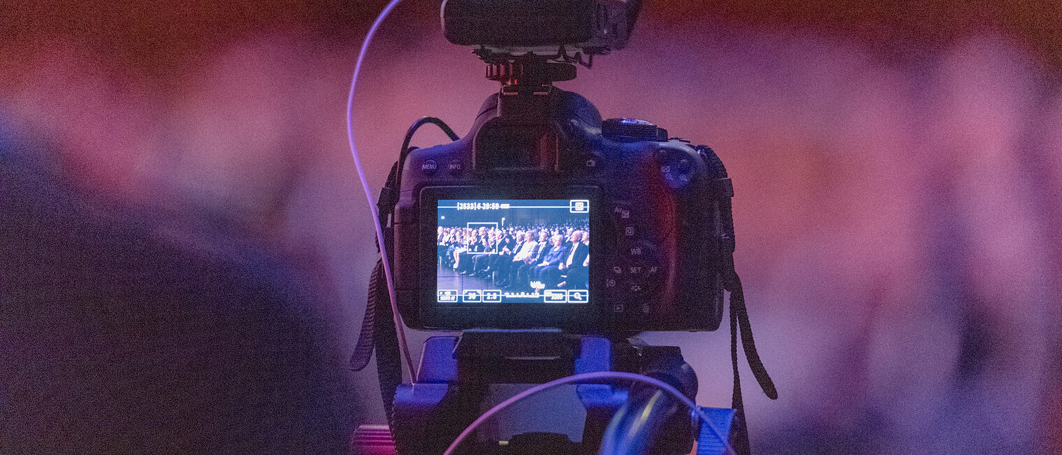 Zu sehen ist ein Fotoapparat und dessen Bildschirm, auf dem wiederum das Publikum der Meisterfeier zu sehen ist. 